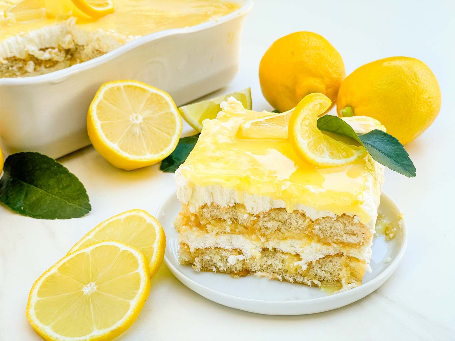 A slice of lemon dessert with cream and lemon slices on top sits on a plate, next to whole and sliced lemons, with a larger dish of the dessert in the background.
