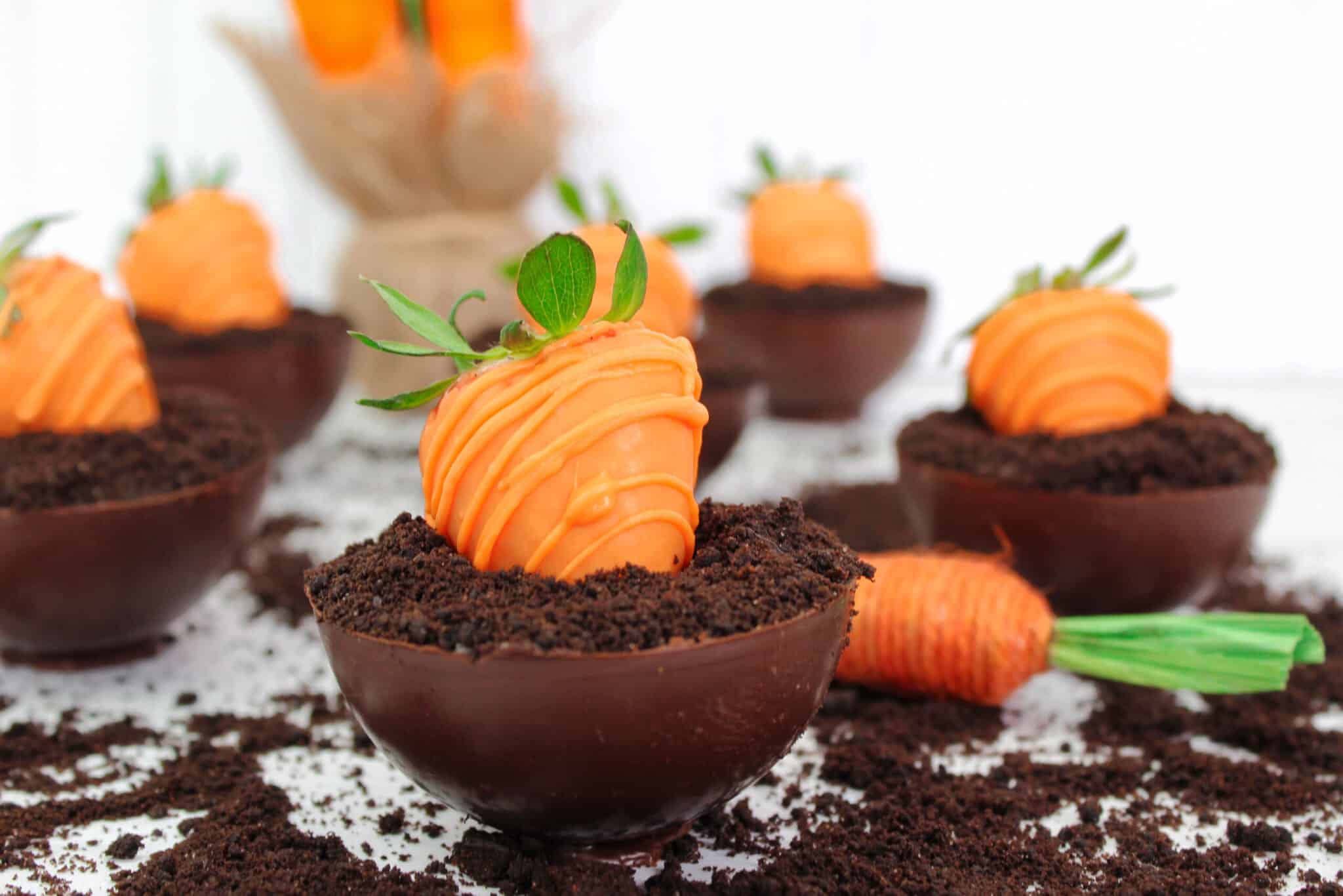 Chocolate bowls filled with crumbled cookies and topped with strawberries dipped in orange coating, resembling carrots planted in soil.