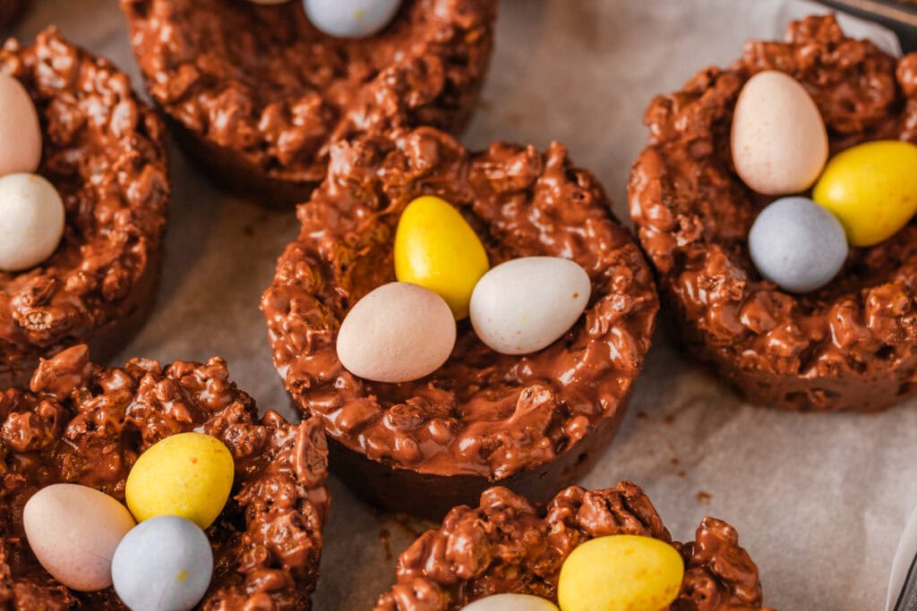 Chocolate rice crispy nests topped with small pastel-colored candy eggs, arranged on a sheet of parchment paper.