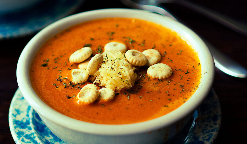 A bowl of tomato soup topped with oyster crackers, shredded cheese, and herbs, served on a blue-patterned saucer with a spoon in the background.