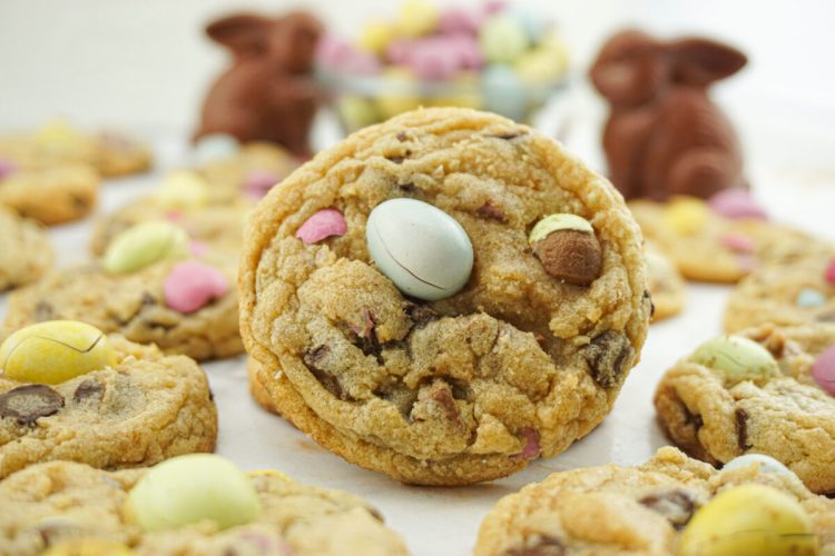 A close-up of a cookie with chocolate chips and pastel candy eggs, surrounded by similar cookies and chocolate bunny figures in the background.