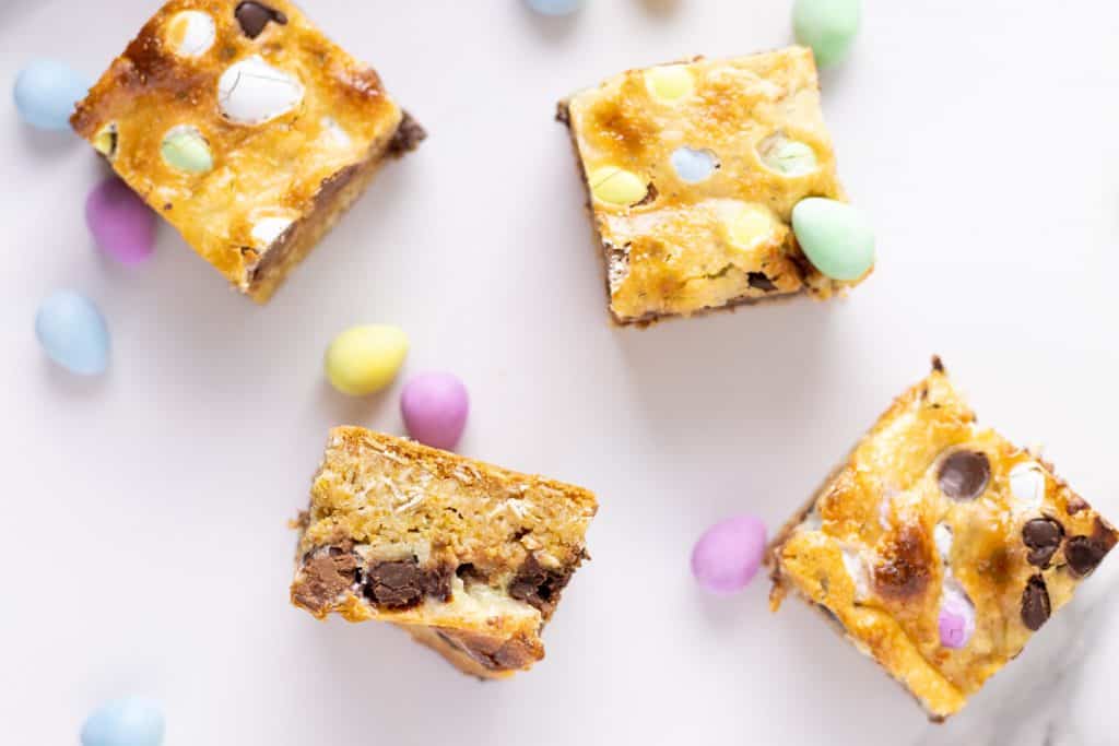 Four blondie bars with chocolate chips and pastel candy-coated chocolates are arranged on a white surface with scattered pastel candies.