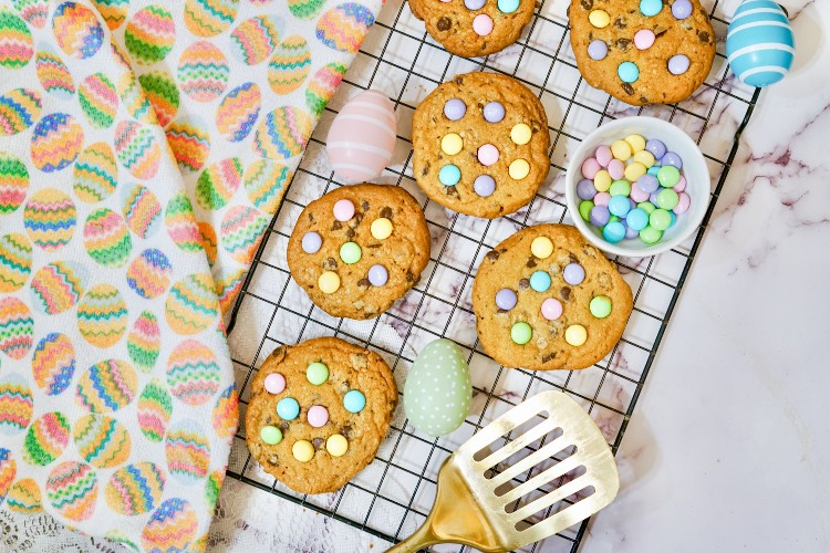 Cookies with colorful candy pieces on a cooling rack, surrounded by pastel Easter eggs, a spatula, and a bowl of candies on a marble surface with a festive cloth.