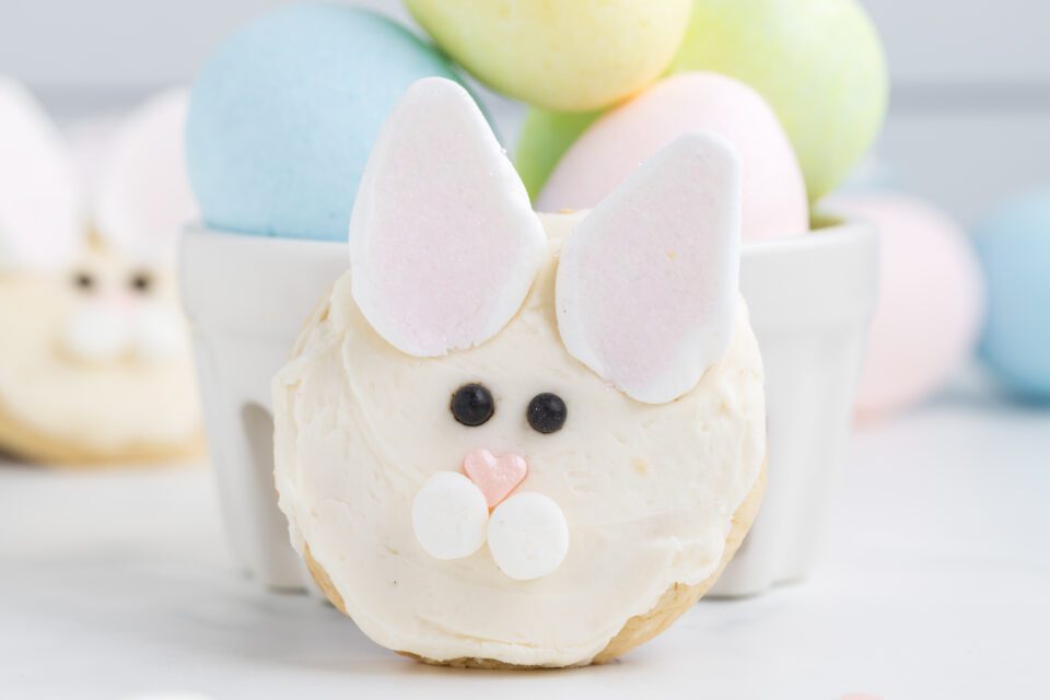 A sugar cookie decorated as a bunny face with white icing, marshmallow ears, candy eyes and nose, with pastel Easter eggs in a white bowl in the background.