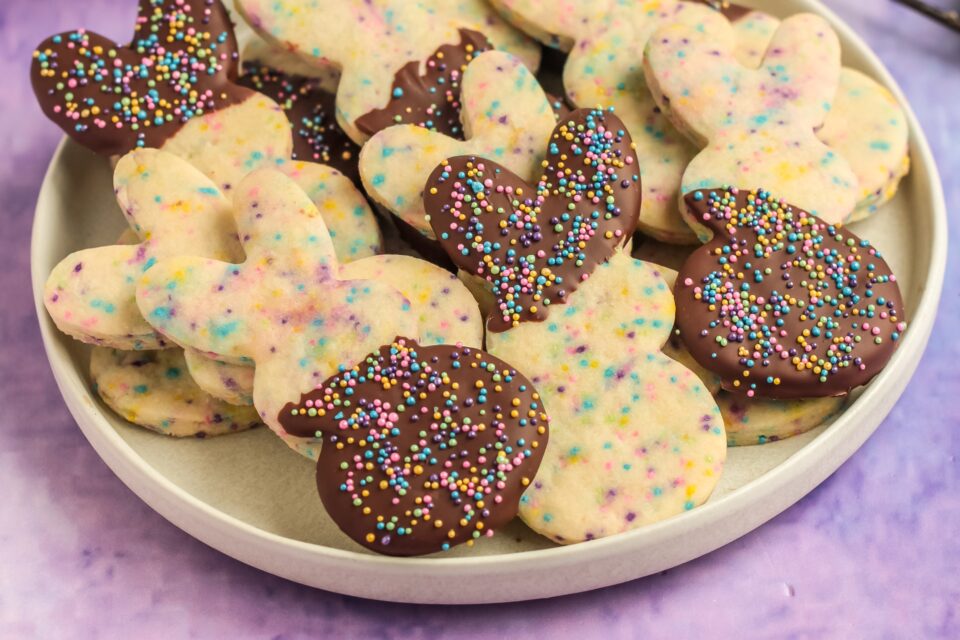 A plate of bunny- and heart-shaped funfetti cookies, some half-dipped in chocolate and topped with colorful sprinkles, on a light purple background.