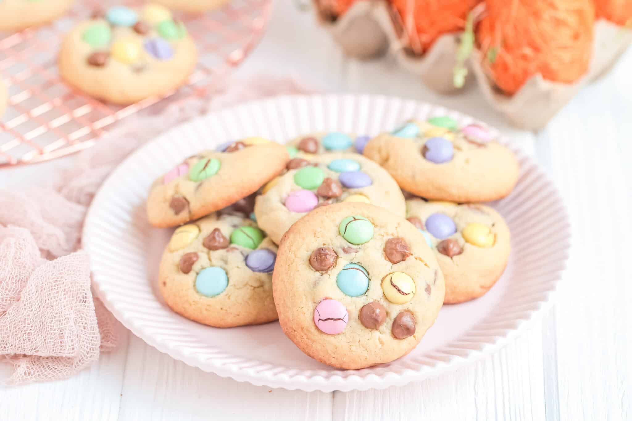 A plate of cookies topped with colorful candy-coated chocolates and chocolate chips, with more cookies and orange decorations in the background.
