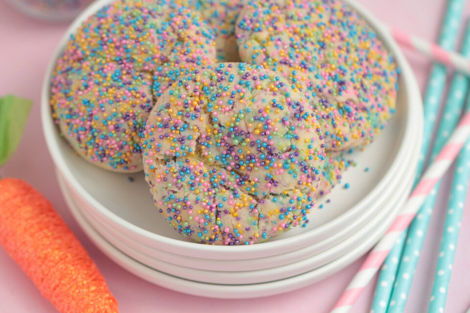 A stack of white plates holds several cookies topped with colorful round sprinkles. Striped straws and a decorative orange carrot are nearby on a pink surface.