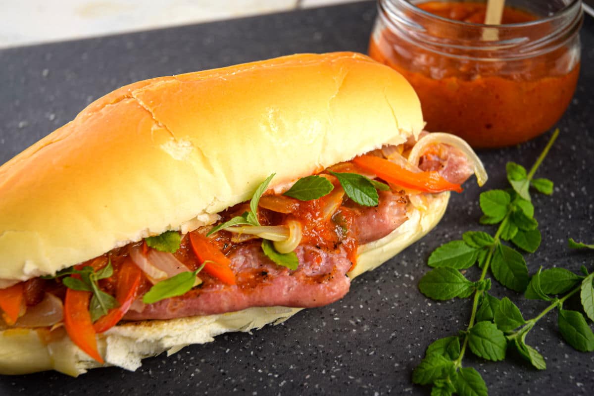 A sausage sandwich with sautéed onions and red peppers on a bun, garnished with fresh herbs, next to a jar of tomato sauce and sprigs of mint on a dark surface.