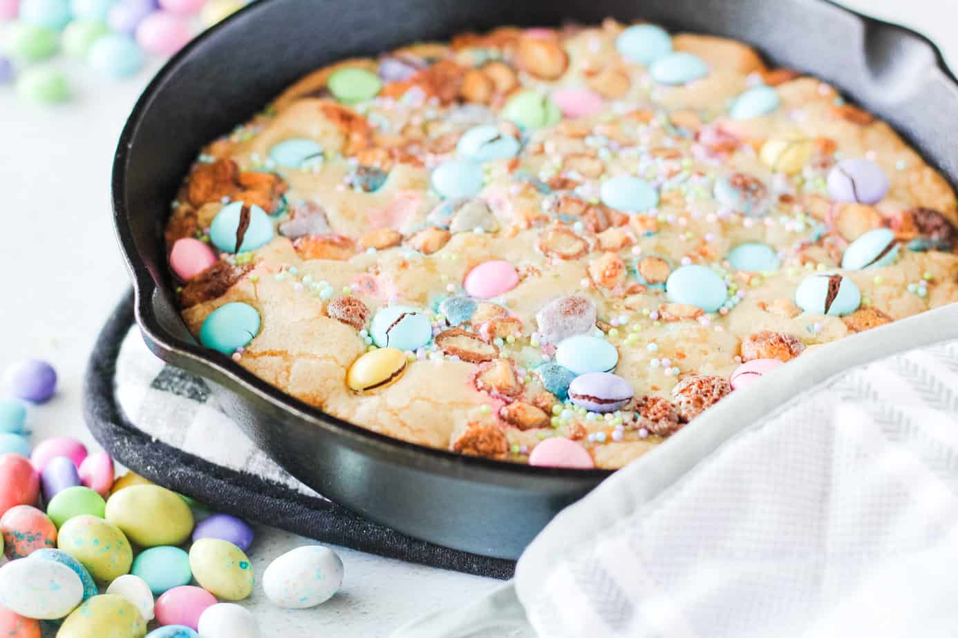 A skillet cookie topped with colorful candy-coated chocolates and sprinkles, sitting on a checkered cloth with scattered candies nearby.