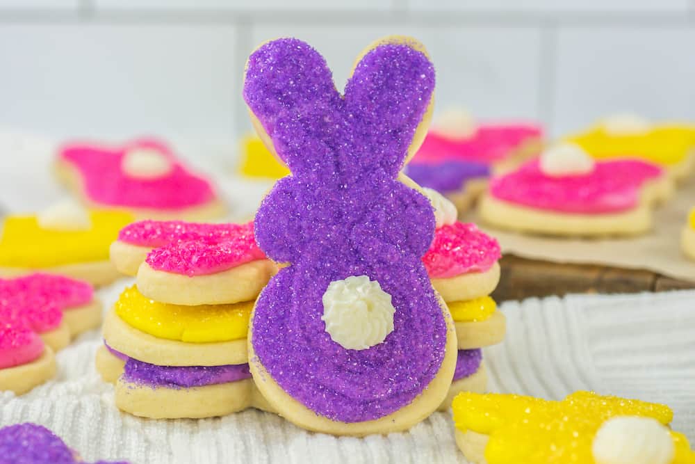 A stack of bunny-shaped sugar cookies decorated with purple, yellow, and pink sugar sprinkles, with white icing for the tails, sits on a white cloth.