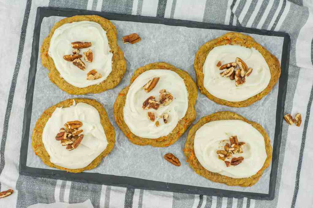 Five frosted cookies topped with pecan pieces are arranged on a parchment-lined tray with a striped cloth underneath.