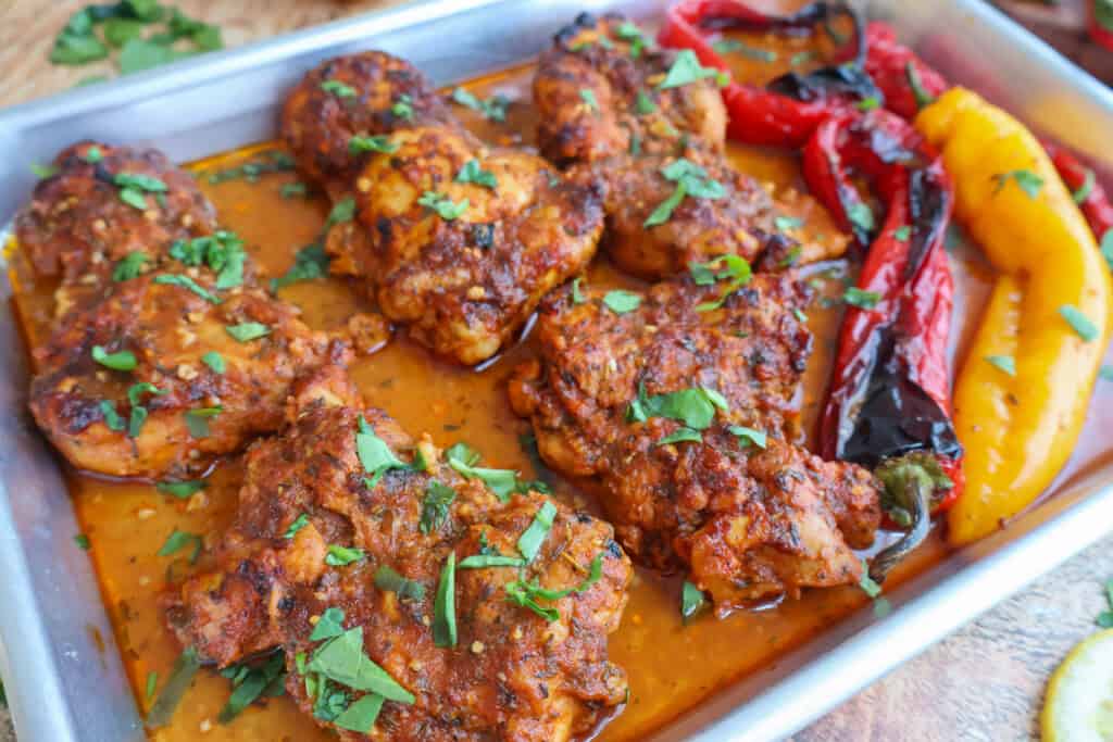 Baked chicken thighs in sauce on a metal tray, garnished with chopped herbs and served with roasted red and yellow peppers.