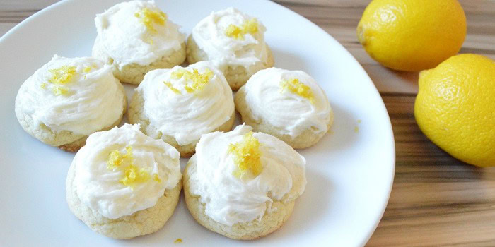 A white plate with seven frosted cookies topped with lemon zest, placed next to two whole lemons on a wooden surface.