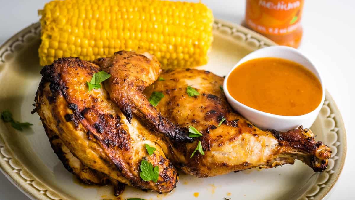 Grilled chicken halves garnished with herbs, served with a corn on the cob and a bowl of orange sauce on a beige plate.