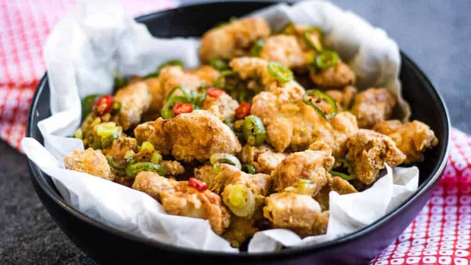 A black bowl lined with parchment paper, filled with pieces of fried chicken garnished with sliced green onions and red chili peppers.