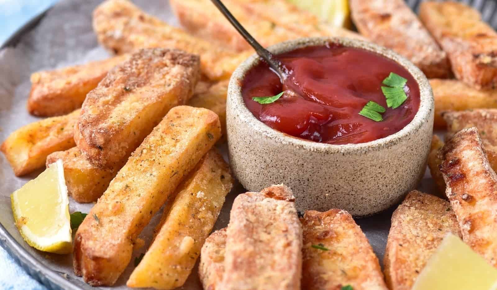 Thick-cut fries arranged around a bowl of ketchup with a spoon, garnished with herbs and lemon wedges on the side.