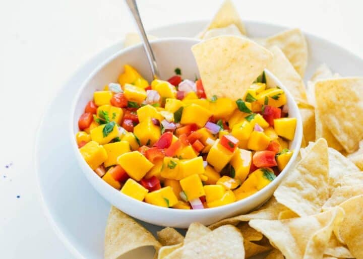 A bowl of mango salsa with diced mango, red onion, and red pepper, garnished with herbs, served with tortilla chips on a white plate.