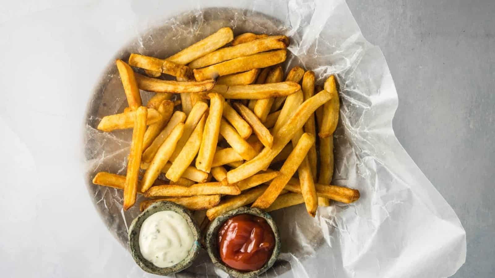 A plate of French fries on parchment paper with two small bowls of dipping sauces, one white and one red, on a gray surface.