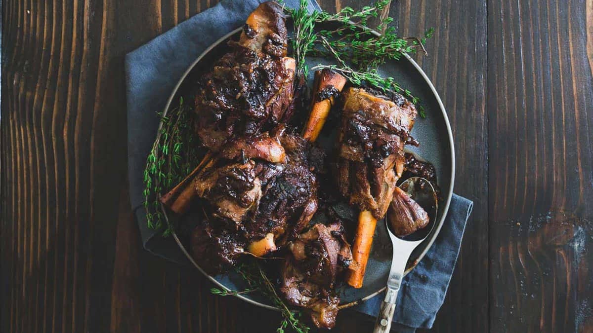 A plate of braised lamb shanks garnished with fresh herbs, served on a dark wooden table with a fork and napkin.