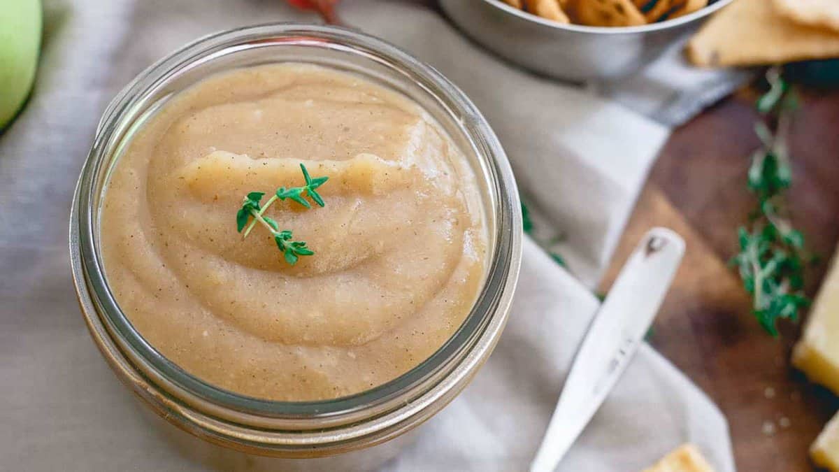 A glass jar filled with creamy applesauce, garnished with fresh thyme sprigs, sits on a table next to a spoon and a bowl of crackers.