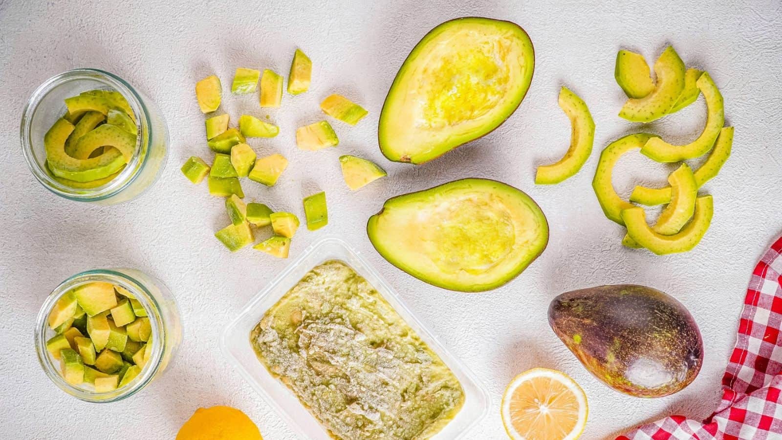 Sliced, diced, and halved avocados with a whole avocado, lemon halves, avocado spread, and a red checkered cloth on a white surface.