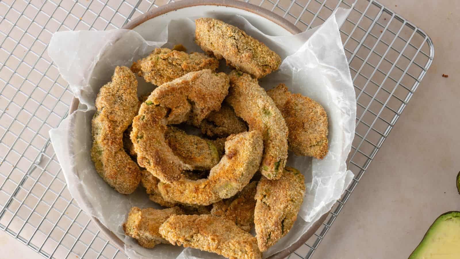 A bowl of breaded and baked avocado slices sits on parchment paper atop a cooling rack.