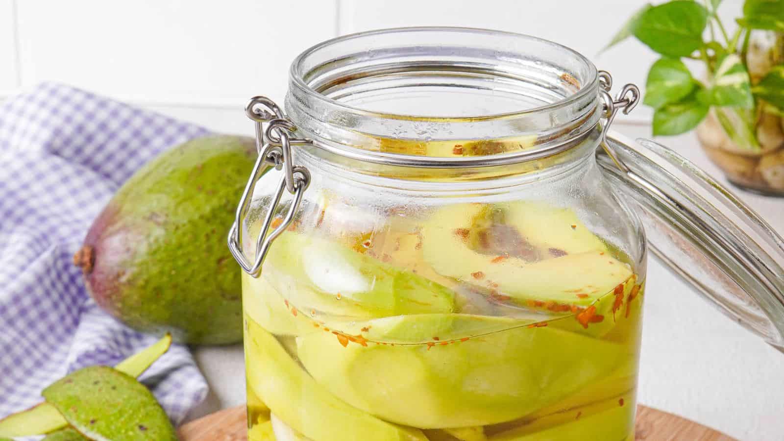 A glass jar filled with sliced green mangoes in brine, with chili flakes visible; an avocado and a checked cloth are in the background.