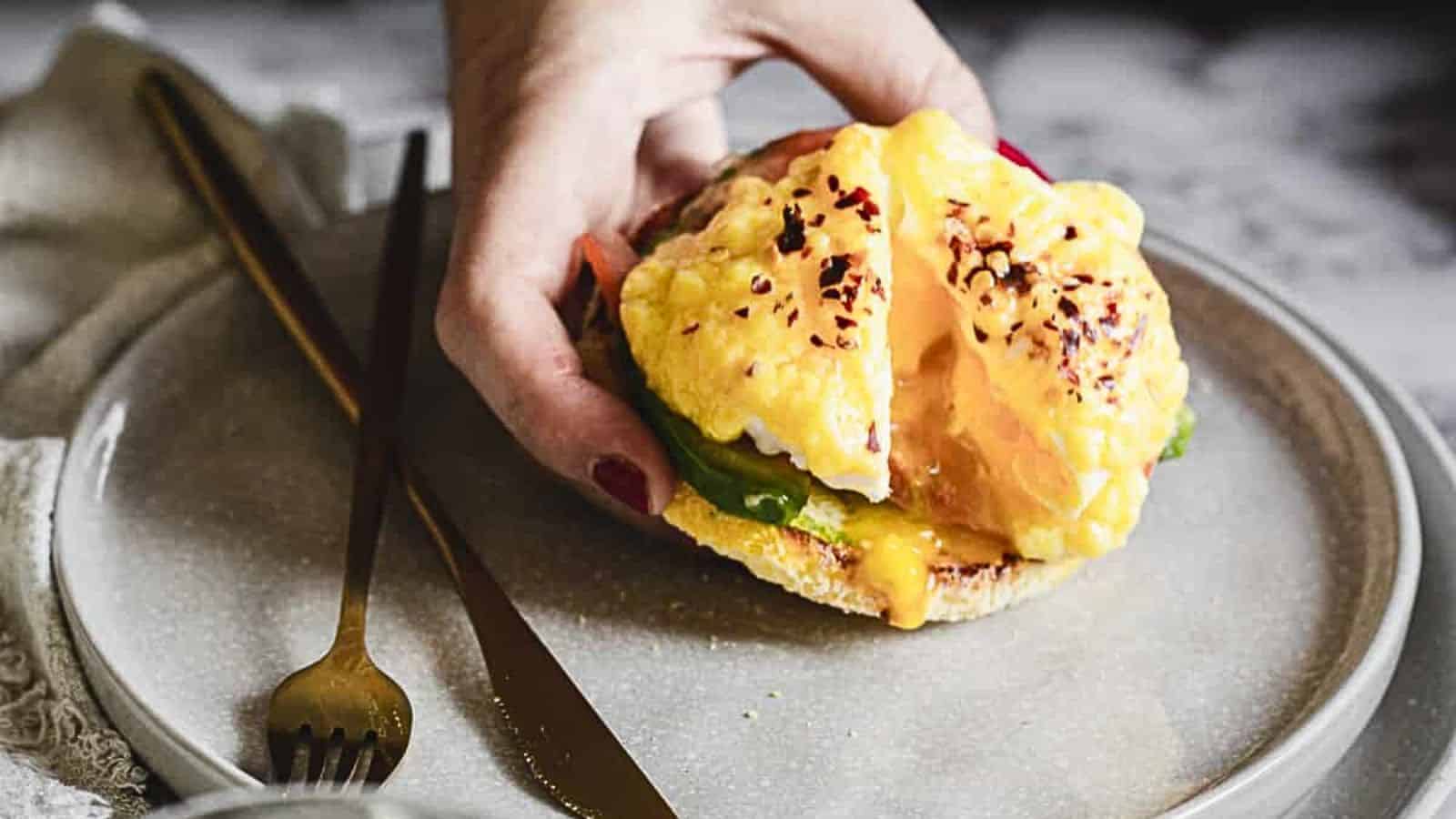 A hand holds half of an English muffin topped with avocado, runny poached eggs, hollandaise sauce, and red pepper flakes on a plate with a fork and knife.