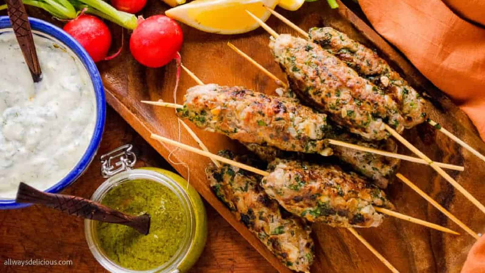 Grilled meat kebabs on wooden skewers served with green sauce, yogurt dip, radishes, green onions, and lemon wedges on a wooden board.