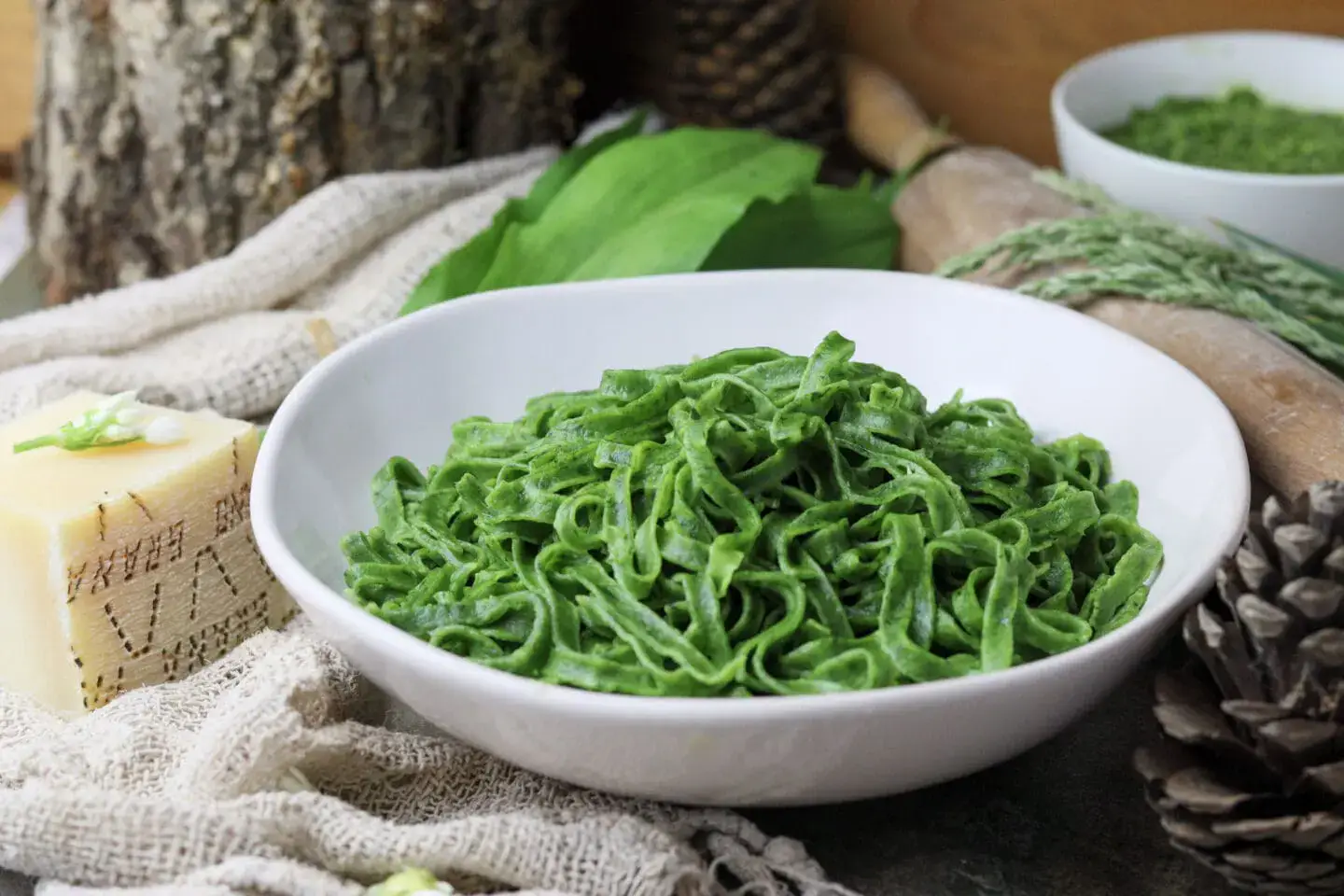 A white bowl filled with green pasta sits on a rustic surface, surrounded by cheese, leaves, a rolling pin, and a small bowl of green sauce.