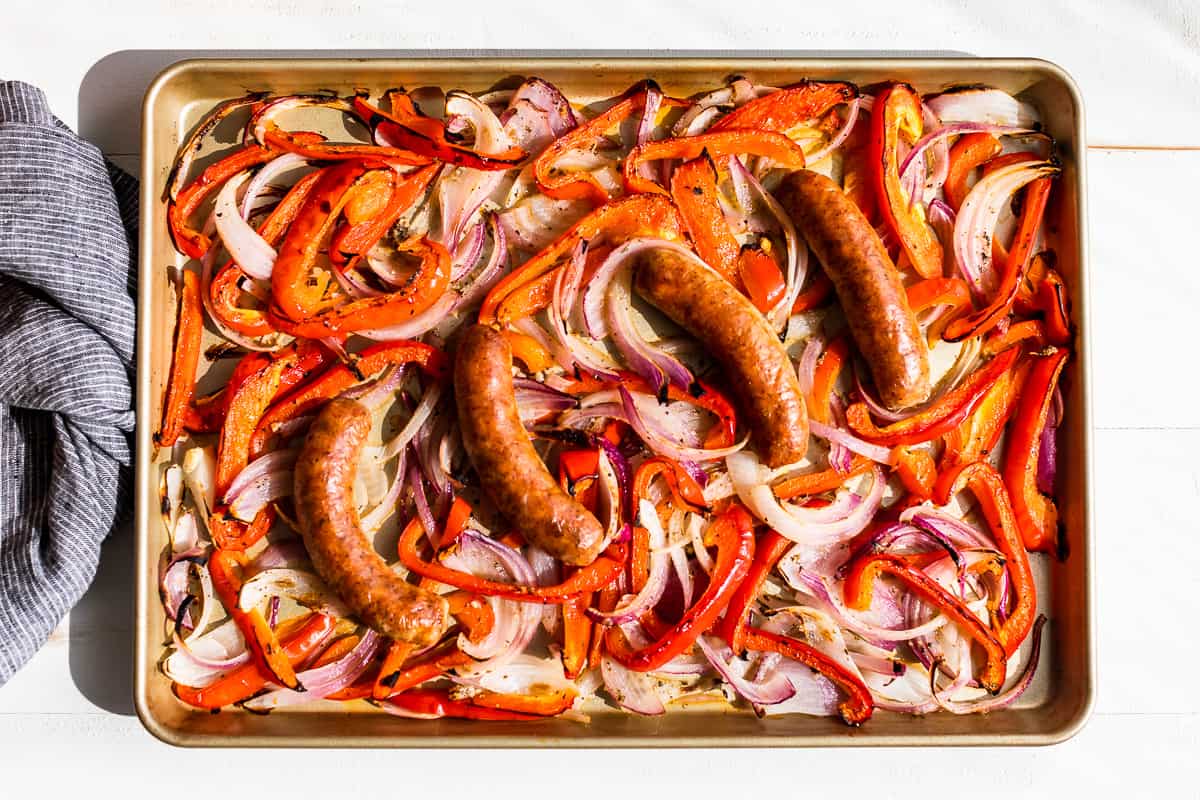 Sheet pan with four cooked sausages on top of roasted red bell peppers and sliced red and white onions. A striped kitchen towel sits to the side.