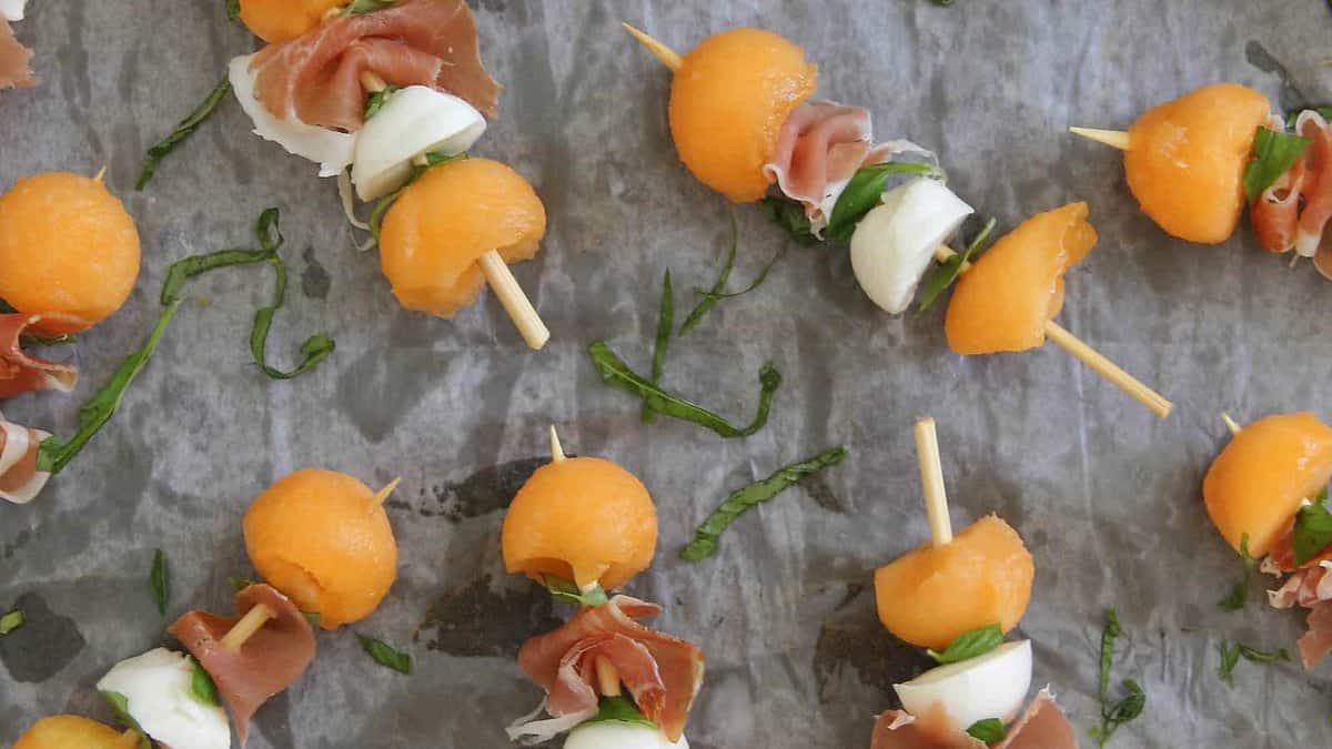 Skewers with cantaloupe balls, mozzarella, prosciutto, and basil leaves arranged on parchment paper.