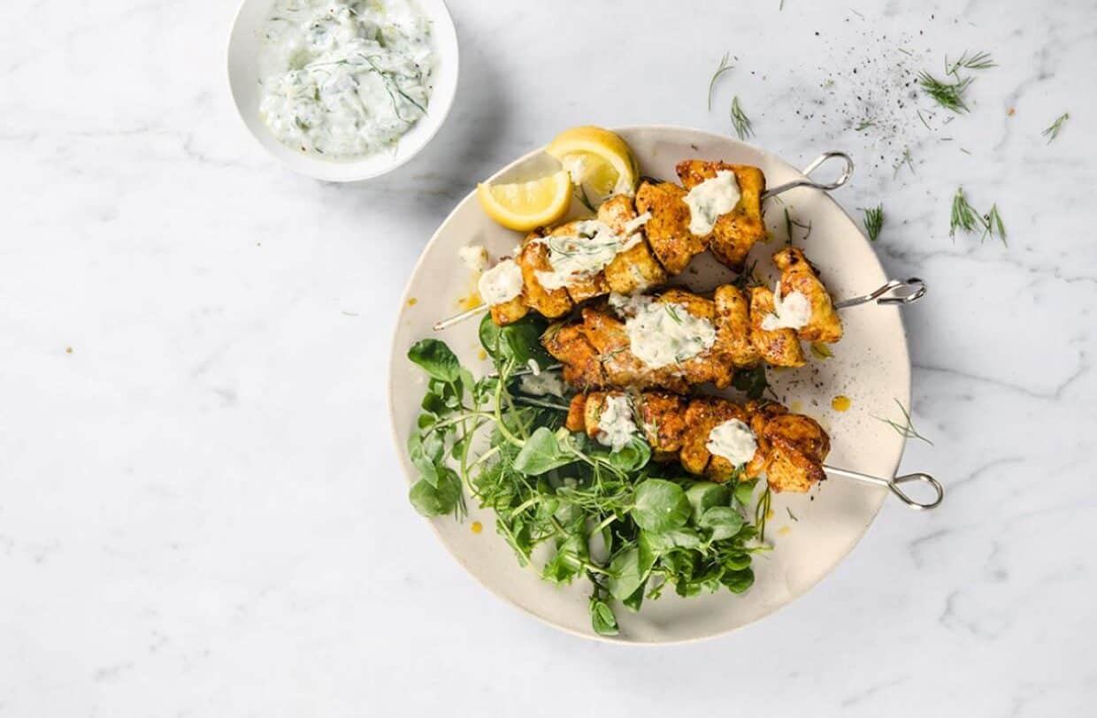 Three grilled chicken skewers with creamy sauce are served on a plate with fresh greens and lemon wedges; a bowl of white sauce is beside the plate.