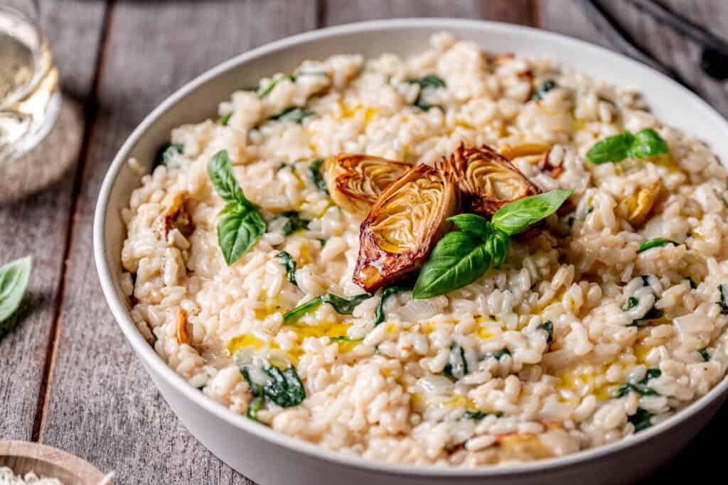 A bowl of creamy risotto garnished with spinach, basil leaves, and roasted artichoke hearts, served on a rustic wooden table.