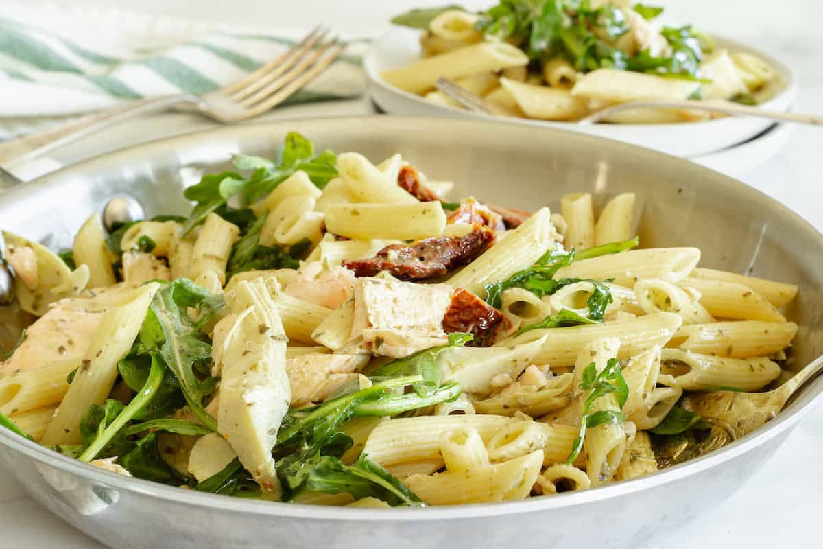 A stainless steel skillet filled with penne pasta, chicken, arugula, and sun-dried tomatoes, with a plate of pasta and a fork in the background.
