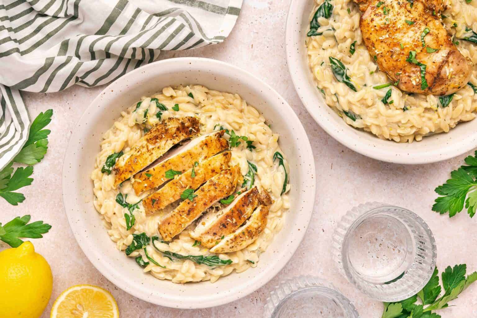 Two bowls of creamy pasta with spinach, each topped with seasoned, sliced grilled chicken; garnished with parsley, next to lemon slices and glasses of water.
