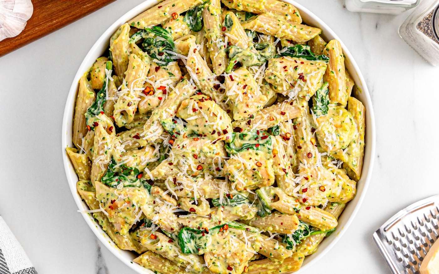 A bowl of penne pasta with spinach, creamy sauce, grated cheese, and red pepper flakes on top, placed on a white surface.