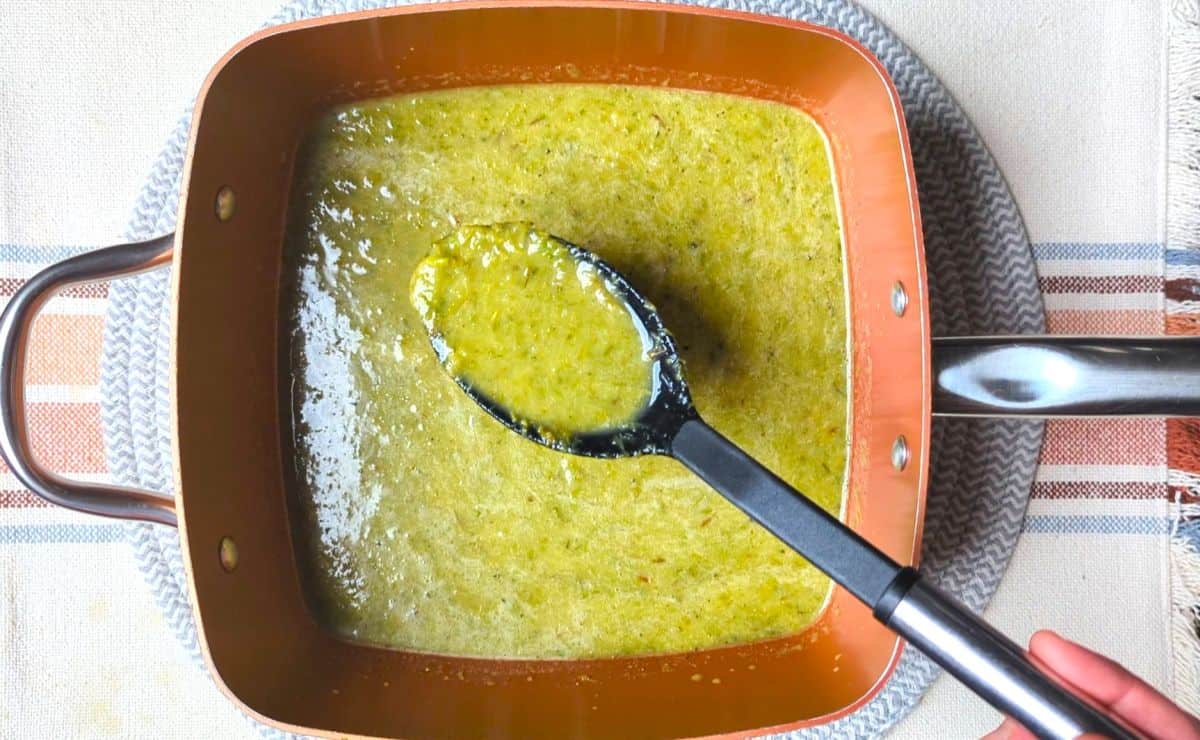 A black ladle lifts up a portion of green soup from a copper pot placed on a woven mat and a striped tablecloth.