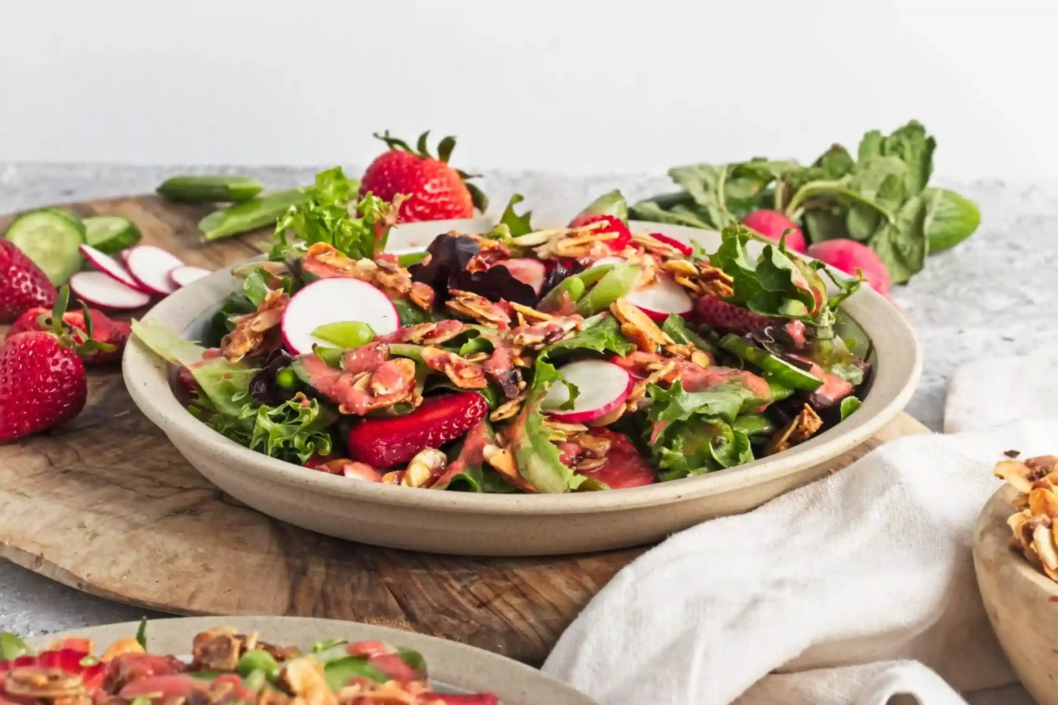 A plate of mixed green salad with sliced radishes, strawberries, and seeds on a wooden surface, surrounded by fresh ingredients.