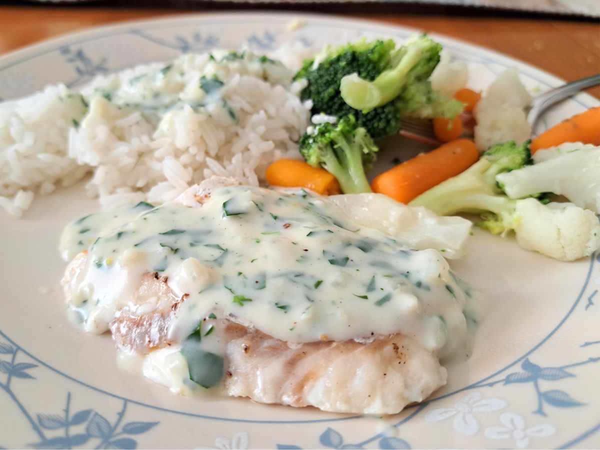 A plate with white fish topped with creamy herb sauce, white rice, and steamed vegetables including broccoli, carrots, and cauliflower.