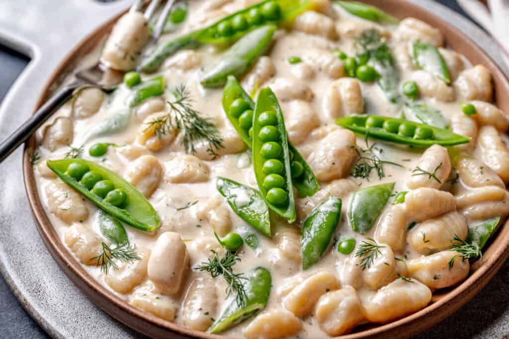 A plate of creamy gnocchi topped with snap peas, green peas, and fresh dill, with a fork resting on the side.