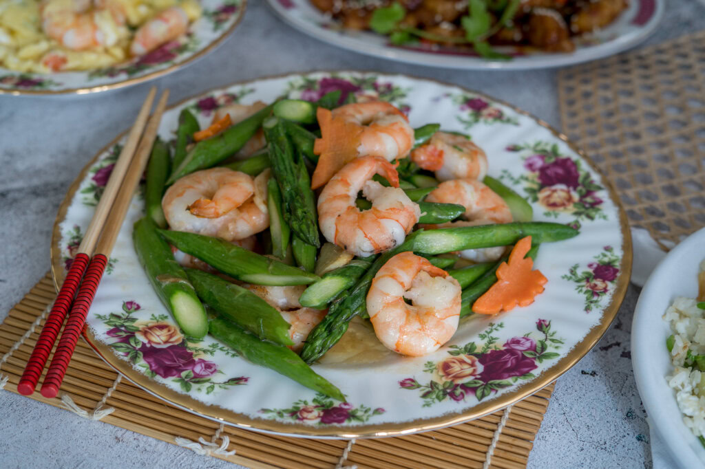 A plate of stir-fried shrimp with asparagus and sliced carrots, served on a floral-patterned dish with red chopsticks resting beside it.