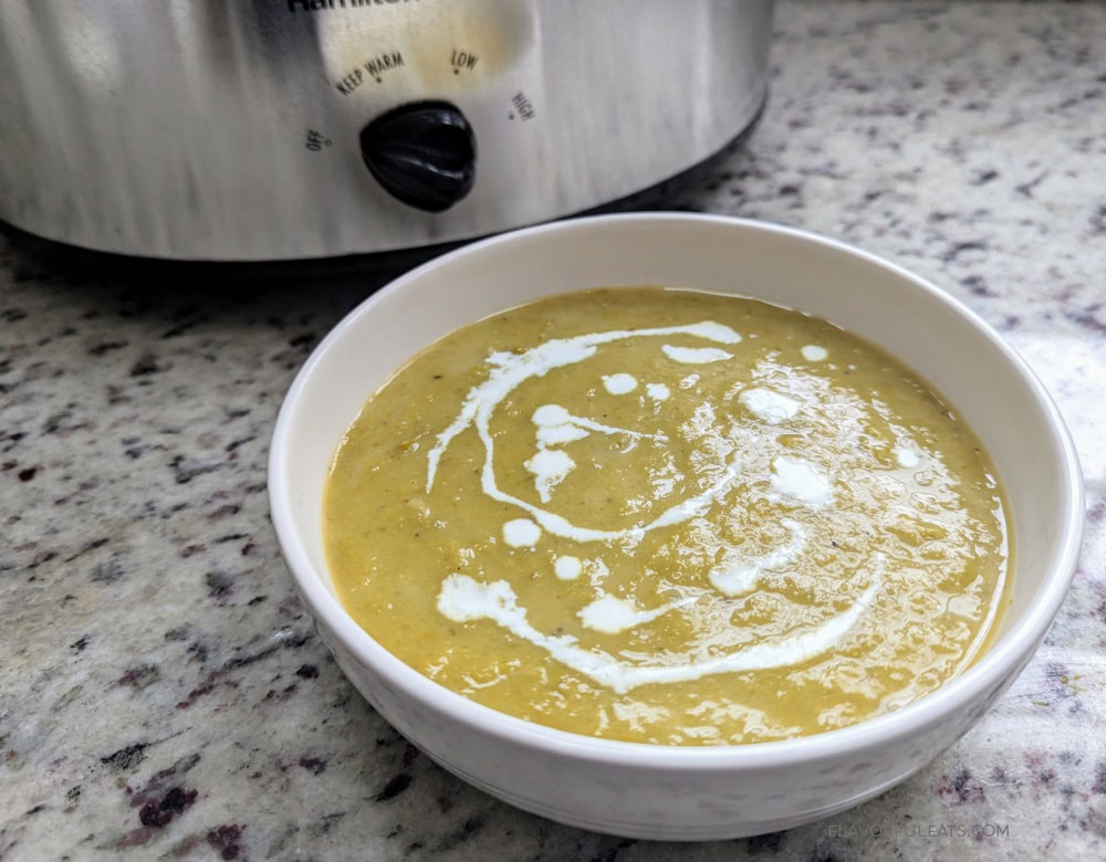 A white bowl of creamy green soup with a swirl of cream on top sits on a granite countertop in front of a slow cooker.