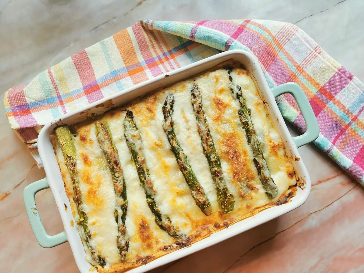 A baked casserole with melted cheese and whole asparagus spears on top, in a light blue dish, placed on a marble counter with a plaid cloth underneath.