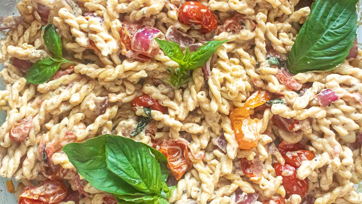 A close-up of pasta mixed with creamy sauce, cherry tomatoes, red onions, and garnished with fresh basil leaves.