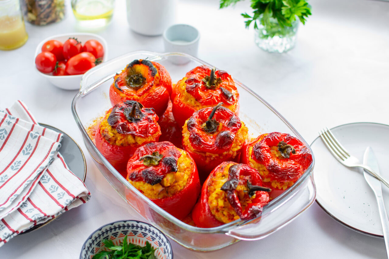 Glass baking dish with six stuffed red bell peppers on a white table, surrounded by plates, cutlery, napkins, and fresh vegetables.