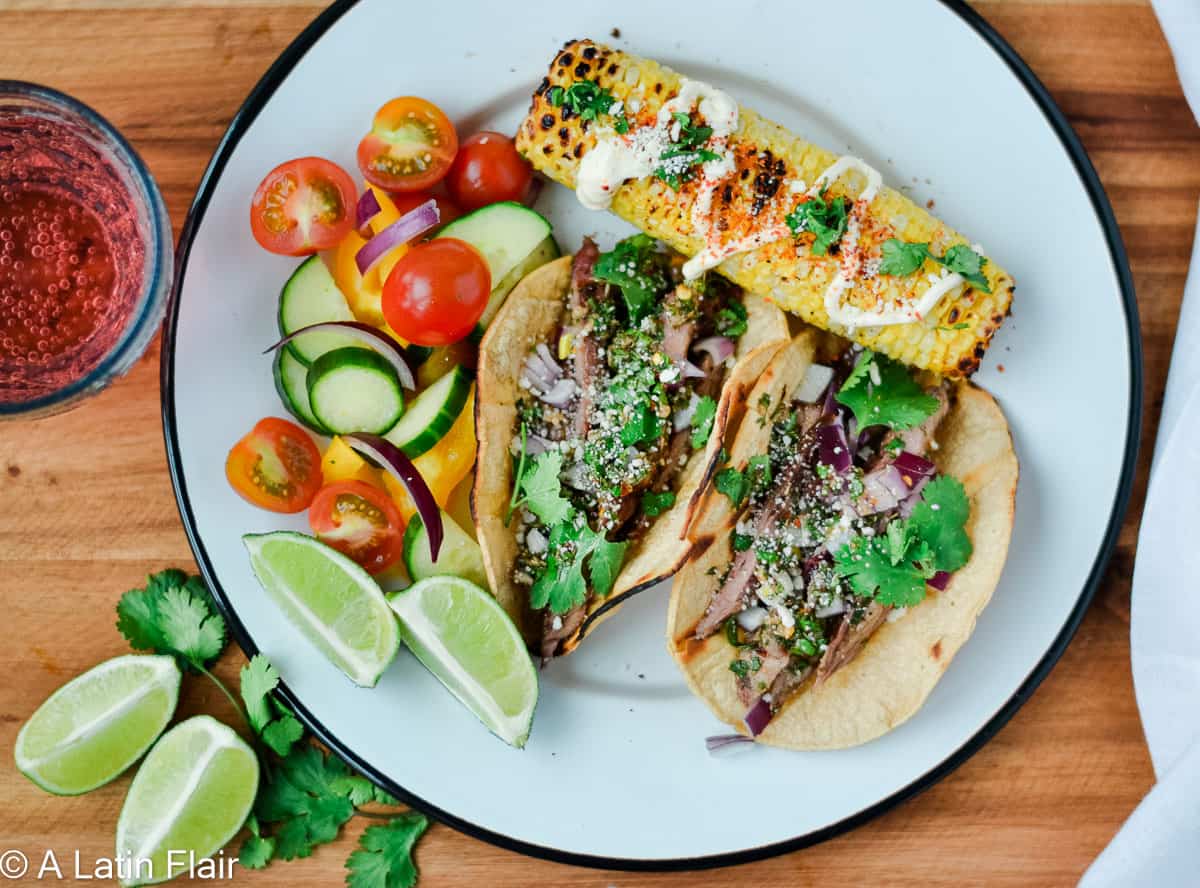 Plate with two tacos topped with cilantro and onions, grilled corn with mayonnaise and spices, a cucumber and tomato salad, lime wedges, and a glass of red drink on the side.