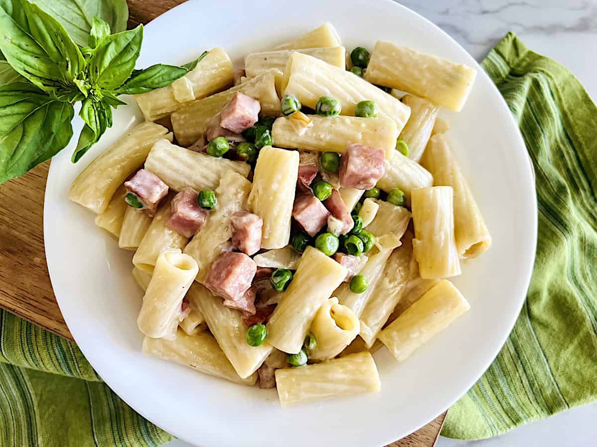 A white plate of rigatoni pasta with diced ham, green peas, and a creamy sauce, garnished with fresh basil on the side.