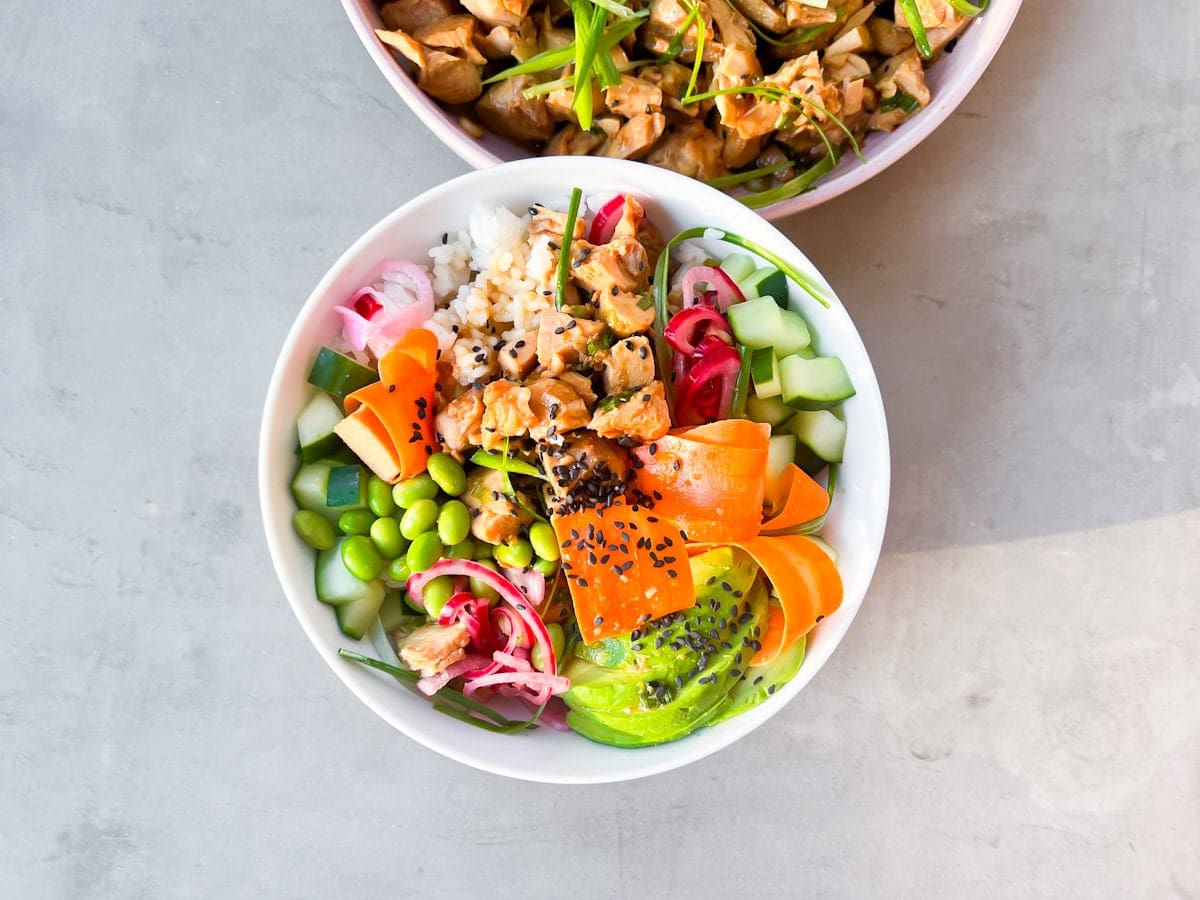 A bowl of rice topped with cubed chicken, sliced cucumber, carrots, avocado, edamame, pickled vegetables, and black sesame seeds, with another bowl partially visible above.