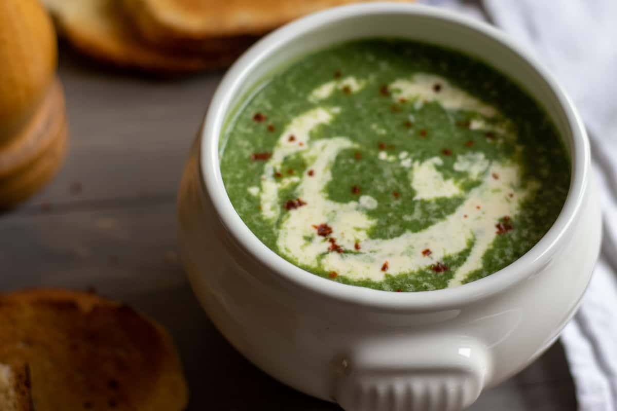 A white bowl filled with creamy green spinach soup, garnished with a swirl of cream and a sprinkle of red pepper flakes. Toasted bread is visible in the background.